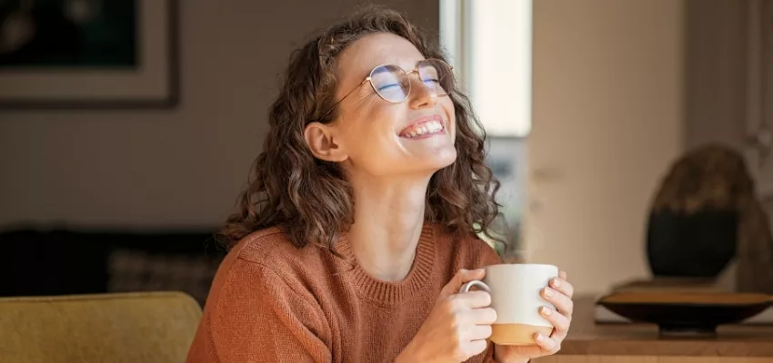 woman wearing sweater sitting on couch holding hot drink and smiling